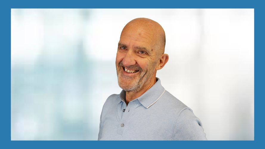 Noel Duffy, smiling at the camera. 
He is wearing a light blue polo shirt. The background is blurred with shades of blue and white, and the image is framed with a dark blue border.